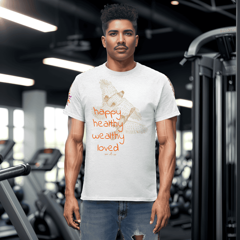 A man stands confidently in a gym, sporting a white T-shirt adorned with vibrant orange text that reads "happy healthy wealthy loved," along with an owl graphic. This Magick "Happy Birthday!" Cotton Tee for Men and Women is an ideal gift choice, set against a backdrop of exercise equipment and large windows.