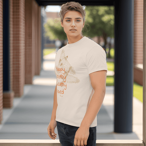 An individual with short hair stands on a walkway bordered by brick columns and greenery, wearing a stylish Magick t-shirt featuring feather illustrations and an inspiring message in orange. This "Happy Birthday!" Cotton Tee for Men and Women is perfect for anyone seeking comfort and motivation.