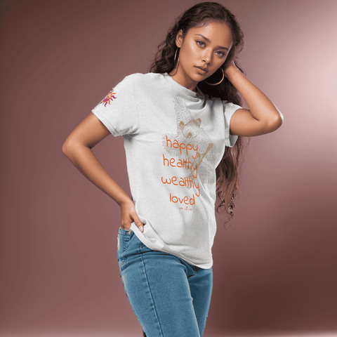 A woman wearing the "Happy Birthday!" Cotton Tee by Magick, featuring the words "happy, healthy, wealthy, loved," poses gracefully against a plain brown backdrop. She complements her outfit with blue jeans and hoop earrings, elegantly standing with one hand on her hip and the other gently touching her hair. This Magick tee is an ideal birthday gift for anyone.