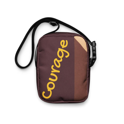 The "Courage" Utility Crossbody Bag by Magick is a small, brown bag with an adjustable black strap, featuring the word "Courage" in yellow script on its front.