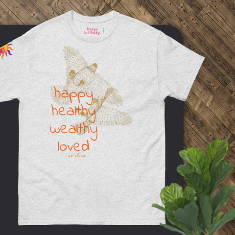 The "Happy Birthday!" cotton tee by Magick features a gray design with a sketch of a koala and the phrase "happy, healthy, wealthy, loved, so it is" in orange text. This delightful shirt is an ideal gift, elegantly displayed on a wooden surface alongside green leaves.