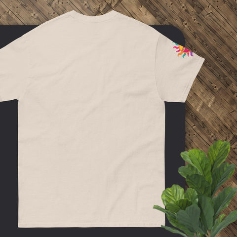 A cream-colored "Happy Birthday!" Cotton Tee by Magick, featuring a vibrant sun design on the right sleeve, is laid flat on a dark surface. The wooden floor can be seen in the background, and there's a partial view of a green potted plant in the corner, making it an ideal gift option.