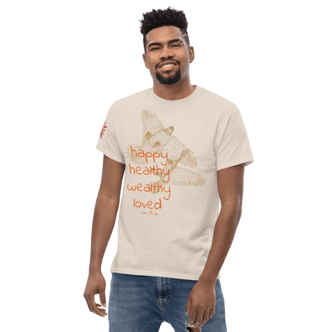 A man wearing the "Happy Birthday!" Cotton Tee by Magick in beige, which features an owl graphic along with "happy, healthy, wealthy, loved" in vibrant orange and red. This T-shirt has a small design on the left sleeve set against a plain backdrop, making it an ideal gift.