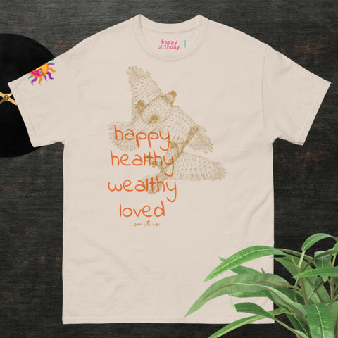 This Magick "Happy Birthday!" Cotton Tee for men and women is a beige, stylish t-shirt showcasing a captivating illustration of an owl in flight, accompanied by the inspirational words "happy, healthy, wealthy, loved." It's an ideal choice for a birthday gift. Displayed on a dark surface with a touch of greenery in the corner.