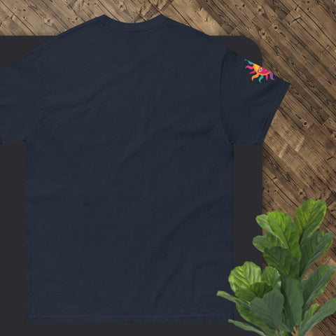 A "Happy Birthday!" Cotton Tee from Magick, in navy blue and designed with a vibrant sun motif on the sleeve, is displayed flat on a wooden floor. A lush green plant peeks into the frame at the bottom right, making it an ideal gift choice.