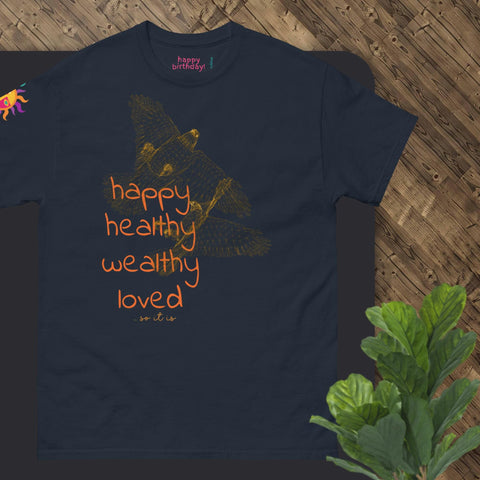 The "Happy Birthday!" Cotton Tee from Magick, available in dark blue, showcases an orange bird graphic accompanied by the phrase "happy, healthy, wealthy, loved" in matching orange text. A vibrant sun adorns the sleeve. This T-shirt makes an ideal birthday gift and is elegantly presented on a wooden surface next to a potted plant.