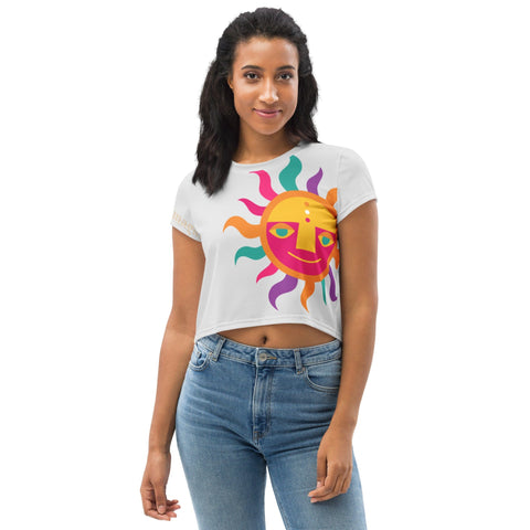 A woman with long dark hair is wearing a stylish wardrobe staple from magickwear: the "Magnificence" Crop Tee, which features a vibrant sun design. She pairs it with blue jeans and stands against a plain white background, smiling gently at the camera.