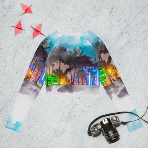 The "Always On" long-sleeve crop top from magickwear, part of our sustainable fashion collection, showcases a vibrant cityscape and palm tree design. It is displayed on a light stone surface alongside two red origami cranes and a vintage camera with a strap.