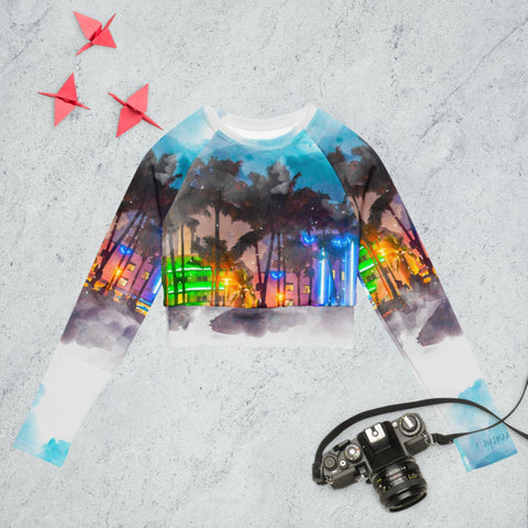 Introducing the "Always On" long-sleeve crop top by magickwear, a stunning blend of style and sustainability. This eco-friendly design features a vivid cityscape with palm trees and neon lights against a textured gray backdrop, offering a chic statement piece. Accompanying this fashionable item is a vintage camera with a strap and two origami cranes, seamlessly merging aesthetic appeal with eco-consciousness.