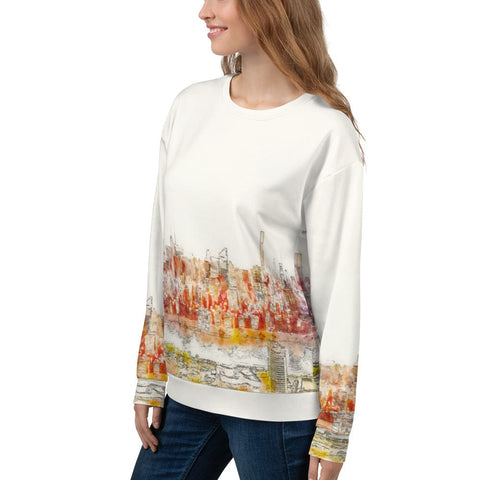 A woman with long brown hair smiles while modeling the stylish and comfortable "Dreamscape" Unisex Sweatshirt from magickwear, which features an all-over print of a colorful abstract cityscape design in shades of red, orange, and yellow at the bottom. She pairs it with blue jeans, standing against a plain white background.