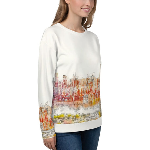 A woman is wearing a stylish and comfortable "Dreamscape" Unisex Sweatshirt by magickwear, featuring an all-over print of a colorful cityscape design along the bottom edge. She is standing sideways, smiling, with her hands relaxed by her sides. She pairs it effortlessly with blue jeans.