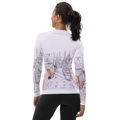 A woman with a ponytail stands with her back facing the camera, wearing the "MY NYC" Women's Rash Guard by magickwear. The white long-sleeve rash guard showcases a detailed sketch of an urban cityscape and offers UPF 50+ Protection. Additionally, it features four-way stretch fabric. She pairs it with black leggings.