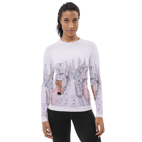 A person with long hair is standing and facing forward. They are wearing a magickwear "MY NYC" Women's Rash Guard, a white long-sleeve shirt featuring a cityscape illustration with buildings and streets. This rash guard boasts UPF 50+ protection, making it perfect for sunny days. The person has a neutral expression and is also wearing black pants.