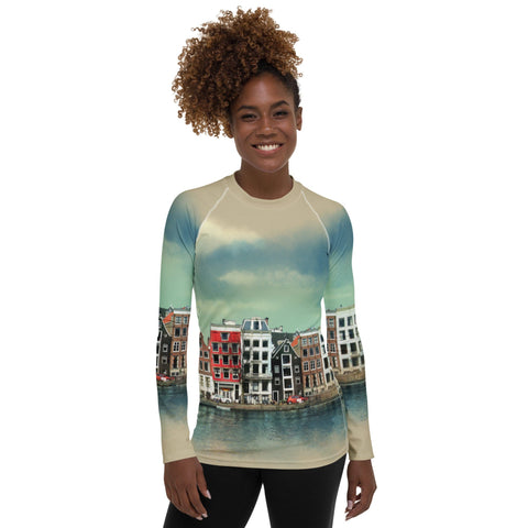 A woman with curly hair smiles while wearing the magickwear "Amsterdam" Rash Guard, which offers UPF 50+ protection and features a vibrant print of buildings along a waterway. The buildings are depicted in various shades of red, yellow, and brown, set against a gradient sky. She stands against a plain background.