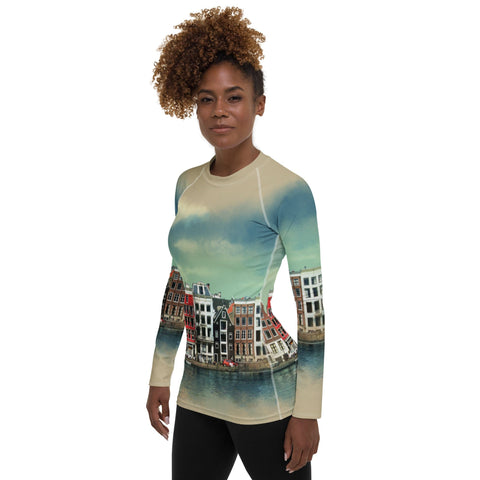 A woman stands facing the camera, showcasing the magickwear "Amsterdam" Rash Guard adorned with a vibrant print of colorful canal houses reflected in water, capturing an Amsterdam cityscape. Crafted from four-way stretch fabric and providing UPF 50+ protection, she complements it with black pants. Her curly hair frames her neutral expression.