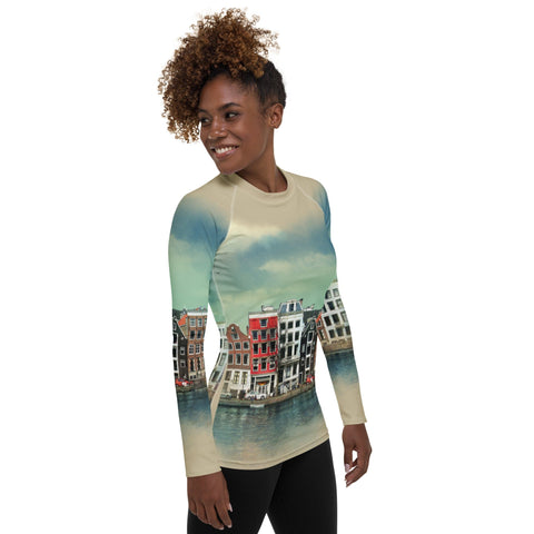 A person with curly hair is wearing a magickwear "Amsterdam" Rash Guard featuring a print of colorful canalside buildings. They are smiling and looking slightly to their left, the four-way stretch fabric ensuring comfort. The background is plain white.