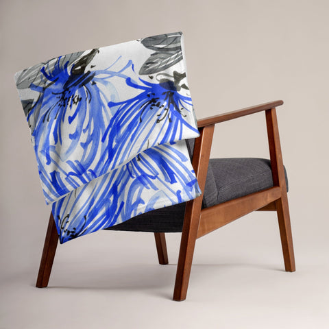 A mid-century modern wooden chair with dark fabric upholstery is placed against a plain background. The chair is complemented by the magickwear Throw Blanket, which features a blue and white floral design and is draped over the backrest, adding a pop of color to the neutral home decor.