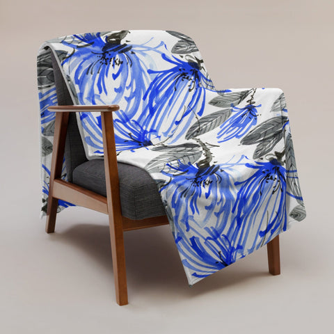 A mid-century modern wooden armchair with a dark gray cushion, draped with the magickwear Throw Blanket, which features a vibrant blue and black floral design on a white background. This elegant piece of home decor is set against a plain neutral backdrop.