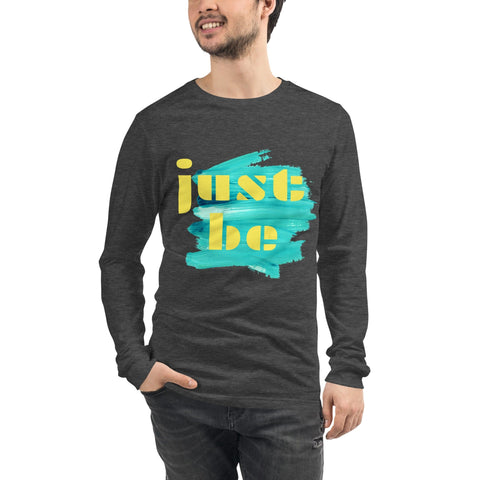 A man is wearing the "Just Be ..." Unisex Lettering Tee by magickwear. This dark gray long-sleeve shirt features a colorful design on the front, with yellow text that reads "just be" over a blue brushstroke. It's comfortable and durable, catering to both men and women. He stands confidently with one hand in his pocket and smiles.