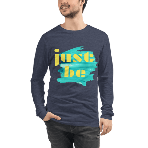 A man wearing a dark blue "Just Be ..." Unisex Lettering Tee from magickwear, featuring bold yellow letters over a turquoise brushstroke design, smiles with one hand in his pocket against a plain background.