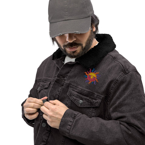 A person wearing the "Snug Warm!" Unisex Denim Sherpa Jacket by magickwear, which features a colorful sun design, adjusts the buttons. With a dark cap and their head slightly lowered, they strike a stylish pose against the plain white background, showcasing the timeless versatility of unisex denim.