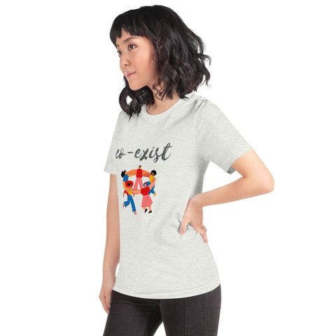 A person with shoulder-length black hair, wearing a stylish light gray "Co-exist" Short-Sleeve Unisex T-Shirt from magickwear featuring the word "co-exist" and an illustration of two people holding hands, stands against a white background. The person has one hand on their hip and is looking to the side.