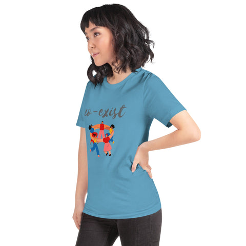 A person with shoulder-length wavy hair is standing sideways, wearing a stylish "Co-exist" Short-Sleeve Unisex T-Shirt by magickwear in blue, which features the word "co-exist" above an illustration of diverse people holding hands around a heart. They have one hand on their hip and are wearing black pants.