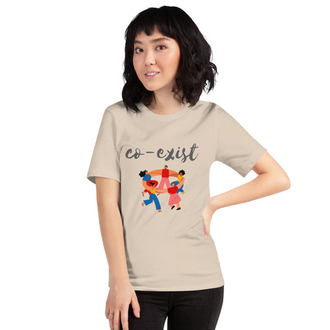 A person with shoulder-length dark hair wears a stylish t-shirt from magickwear that says "co-exist" in cursive. The beige, short-sleeve unisex t-shirt, named "Co-exist," features an illustration of diverse individuals holding hands and dancing in a circle. The person stands against a plain white background, smiling slightly.