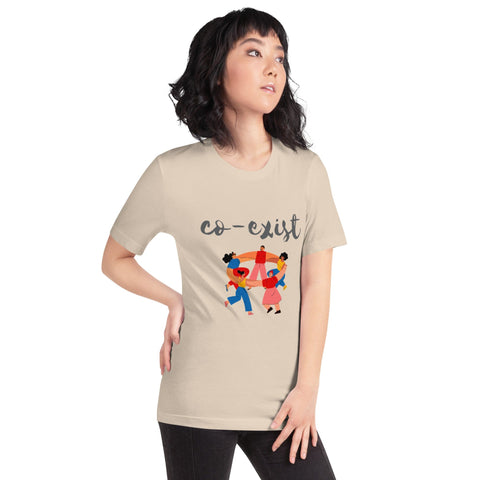 A person with shoulder-length black hair stands wearing a stylish beige "Co-exist" Short-Sleeve Unisex T-Shirt from magickwear, featuring the word "co-exist" above an illustration of diverse people holding hands in a circle. The person gazes off to the side with one hand on their hip.