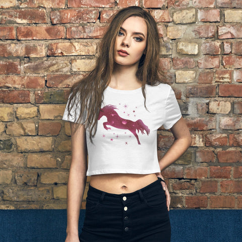 A person with long hair stands against a brick wall, wearing the stylish "M Moment" Women’s Crop Tee by magickwear, which features a pink horse design with stars. They have one hand in the pocket of their black pants, looking directly at the camera to showcase their chic style and the slim fit of this trendy garment.