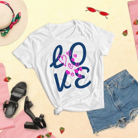 Flat lay of a summer outfit featuring magickwear's "LOVE Big" Women's Short Sleeves Lettering T-shirt in white, paired with denim shorts, black sandals, a floral-banded sun hat, and red sunglasses on a sandy background sprinkled with strawberries.